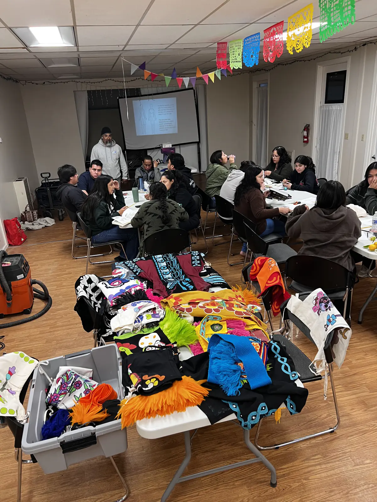 Members gather indoors for a regalia workshop, papel picado banners overhead